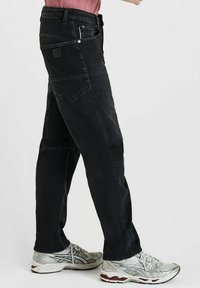 Persoon in losse zwarte jeans met grote zijzakken en zilverkleurige hardloopschoenen, staande tegen een effen lichte achtergrond.