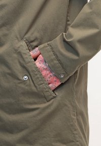 Chaqueta verde oliva con un bolsillo lateral, que cuenta con un cierre de botón a presión. La manga muestra un tatuaje rosa con manchas oscuras. Tela suave y texturizada.