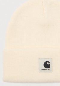 Cream-colored ribbed knit beanie with folded cuff and a square white patch displaying a black Carhartt logo.