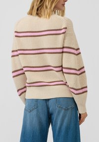 Beżowy sweter na drutach z różowo-brązowymi poziomymi paskami, obniżonymi ramionami i prążkowanym dołem. Noszony z niebieskimi jeansami o szerokich nogawkach.