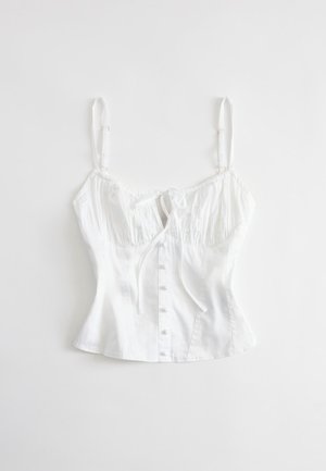 Blouse blanche sans manches ajustée avec bretelles réglables, boutons à l'avant et détail d'un nœud noué au niveau de la poitrine, posée à plat sur un fond blanc.
