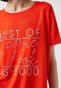 Czerwona koszulka z krótkim rękawem z błyszczącymi literami z srebrnych cyrkonii, na której widnieje napis "BEST OF New York JS/2000", o miękkiej, gładkiej fakturze.