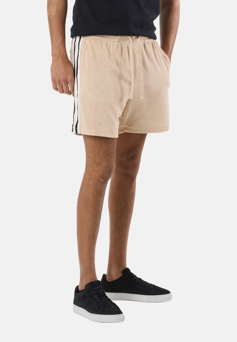 Homme portant un short en tissu éponge beige avec des bandes latérales blanches et noires, un t-shirt noir et des baskets noires avec des semelles blanches.