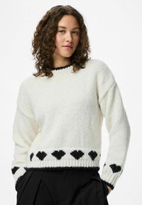 Noor naine lokkis juustega, kes kannab valget sviitrit, mille ääres ja varrukatel on mustad südame muster, koos mustade pükstega.