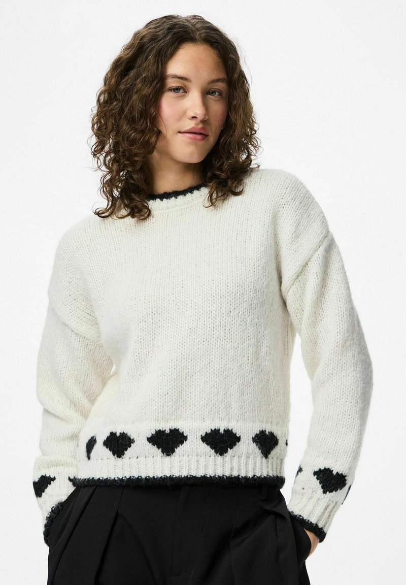 Noor naine lokkis juustega, kes kannab valget sviitrit, mille ääres ja varrukatel on mustad südame muster, koos mustade pükstega.