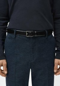 Mann trägt dunkelblaue Hose mit dezitem Karomuster, schwarzen Ledergürtel und schwarzes Langarmhemd, Hände entspannt an den Seiten.