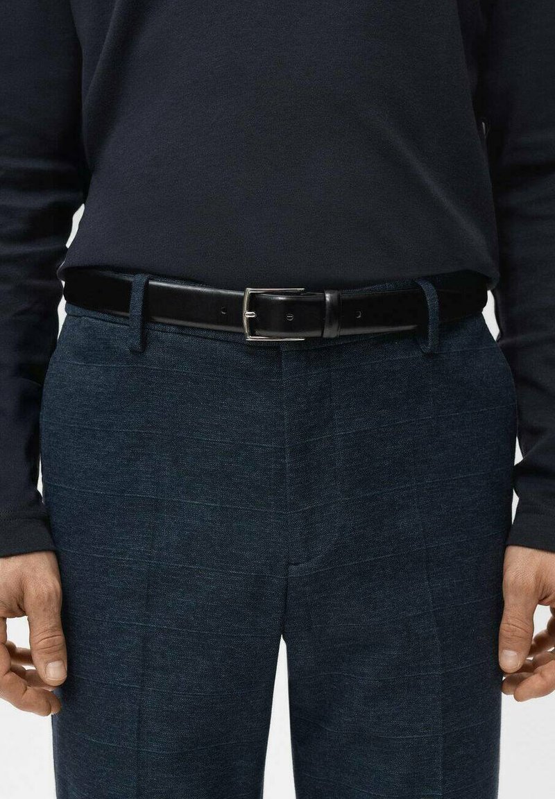 Homme portant un pantalon bleu marine foncé à motif à carreaux subtil, ceinture en cuir noire, et chemise noire à manches longues, les mains détendues le long du corps.