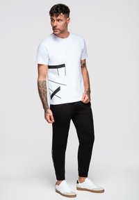 Biała koszulka z czarnym wzorem graficznym, w połączeniu z czarnymi spodniami dresowymi i białymi trampkami z brązowymi akcentami. Widać tatuaże na ramionach.