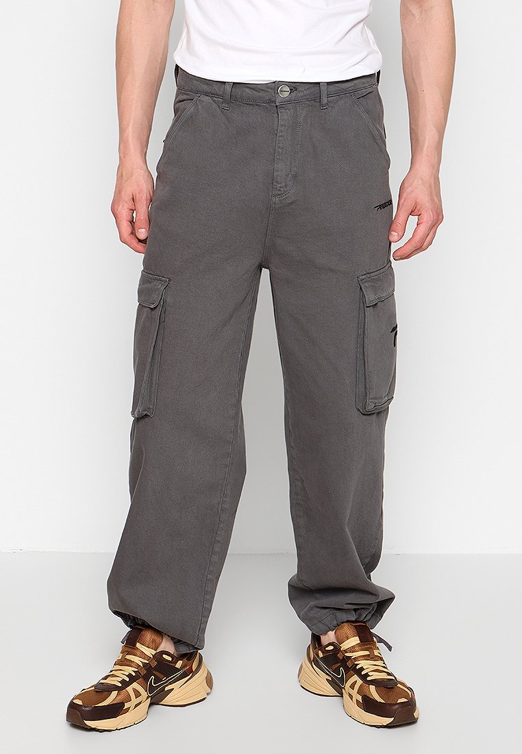 Pantalon cargo gris à coupe décontractée, doté de poches latérales, d'une fermeture par bouton et de détails de couture visibles. Porté avec des baskets à motifs.