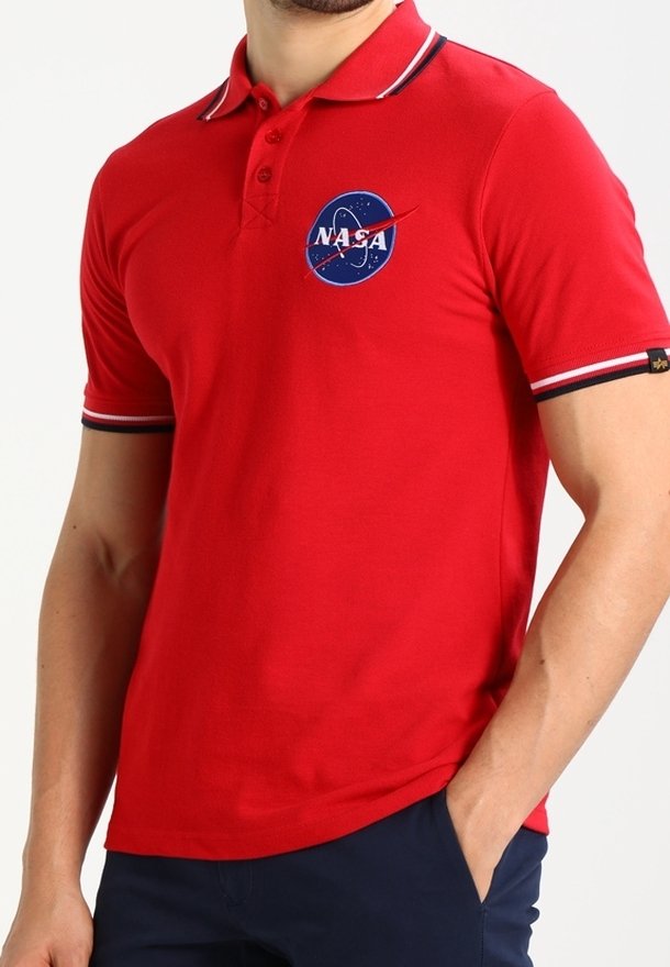 Homme portant un polo rouge NASA avec un liseré rayé blanc et noir sur les manches et le col, main dans la poche d'un pantalon bleu marine.