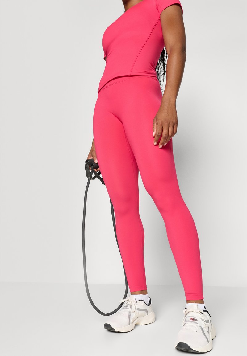 Strakke sporttop en leggings in felroze, gemaakt van rekbaar materiaal. In één hand wordt een zwarte springtouw vastgehouden. Witte sneakers met zwarte accenten.