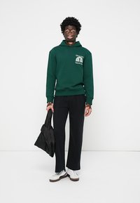 Grøn hættebluse med en frontlomme og logo; parret med sorte bukser og hvide sneakers. Holder en sort taske.