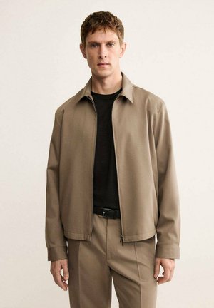 Giacca zip-up color marrone chiaro realizzata in tessuto liscio, con colletto classico e tasche laterali, indossata sopra una maglietta nera e pantaloni coordinati.