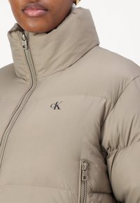 Giacca puffer beige con colletto alto, chiusura con zip e dettagli con logo. Il materiale sembra morbido con una texture trapuntata e tasche con zip.