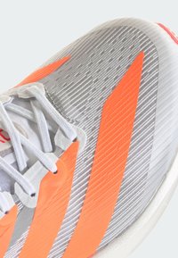 Scarpa da ginnastica grigia con tomaia strutturata. Presenta strisce arancioni brillanti, lacci grigio chiaro e suola bianca.