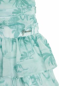 Robe vert menthe avec un motif tropical de palmiers, détails en volants superposés et un accent logo argent "Guess". Tissu léger et fluide.