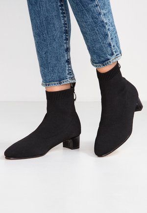 Person iført sorte strikkede sokkestøvler med lave blokhæle og blå opkørte jeans, stående på en lys gulv.