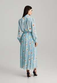 Robe bleu clair avec un motif floral, manches longues, taille élastique et jupe fluide. Portée avec des chaussures à talons hauts noires.