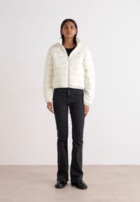 Hvit pufferjakke med en cropped design, med horisontale seksjoner og glidelås foran. Kombinert med svarte vide jeans og svarte sko.