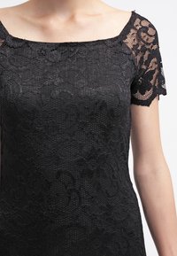 Černý krajkový top se čtvercovým výstřihem, krátkými rukávy a složitými květinovými vzory, s texturovaným povrchem a průsvitnými akcenty.