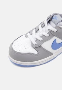 Sneaker grigio e bianco con tomaia in pelle texturizzata, dettagli blu e punta perforata. Presenta lacci bianchi e suola in gomma.