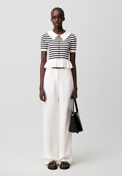 Mujer con top de manga corta a rayas blancas y negras con bajo fruncido, pantalones anchos blancos, bolso negro y sandalias oscuras, de pie frente a un fondo gris.