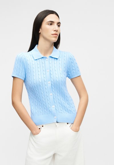 Sieviete valkā gaiši zilu īsroku kabeļadījuma pogājamu džemperi ar apkakli un baltas bikses, rokas kabatās, skatās uz sāniem.