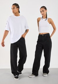 Svart bredbeinte jeans, hvit oversized t-skjorte, og hvit topp. Modeller har på seg svarte og hvite sjakkrutete sneakers. Glatte teksturer, avslappet stil.