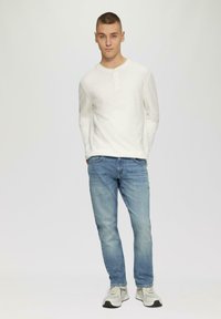Chemise henley à manches longues blanche avec trois boutons, associée à un jean bleu clair et des chaussures de sport grises, se tenant devant un fond uni.