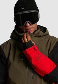 Gant imperméable rouge avec des accents noirs, tissu texturé, fermeture Velcro et détail de logo, porté par-dessus une veste verte et un bonnet noir.