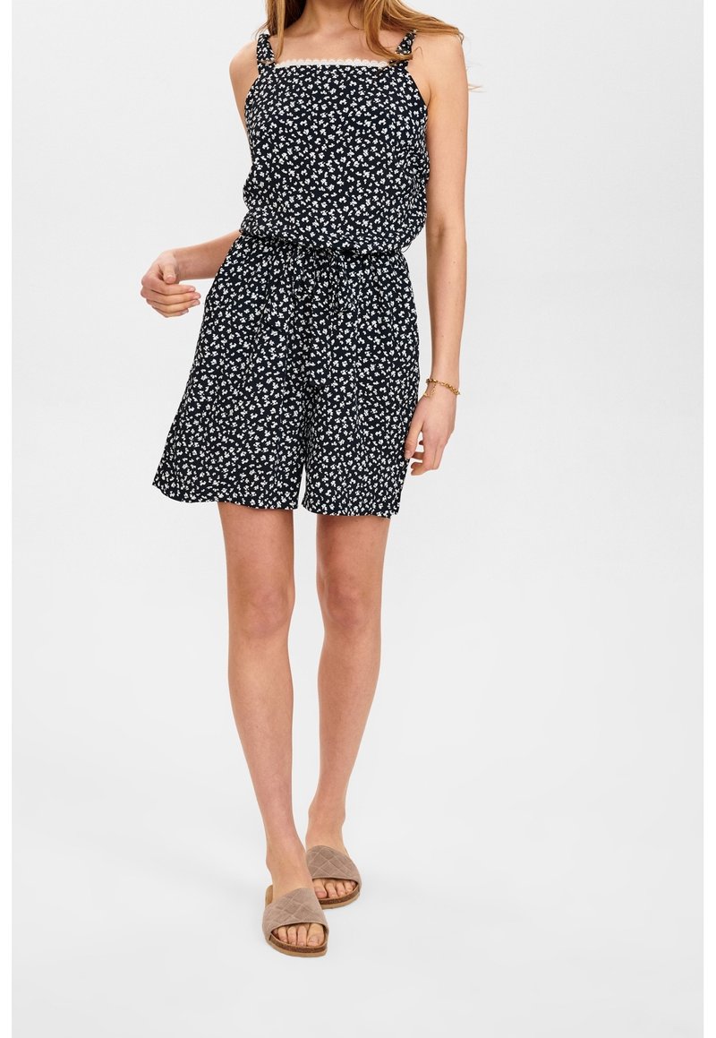 Žena oblečená v černém květinovém overal s odhalenými rameny a zavázaným pasem, na béžových pantoflích, stojící na bílém pozadí.