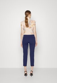 Blommig volangtop i persika med lila och gula blommor, tillsammans med marinblå smalbyxor. Modellen bär svarta klacksandaler.