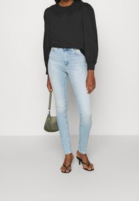 Svart blus med en dekorativ krage, ljusblå slim-fit jeans, svarta remsandaler och en liten olivgrön handväska.