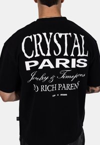 Černé bavlněné tričko s výrazným bílým textem na zadní straně, včetně "CRYSTAL PARIS" a "ŽÁDNÍ BOHATÍ RODIČE." Krátké rukávy, volný střih.
