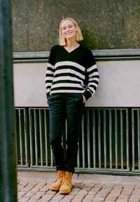 Fekete-fehér csíkos kötött pulóver V kivágással, fekete nadrággal és világosbarna fűzős csizmával, egy texturált fal mellett állva.