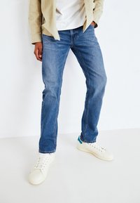 Lichtblauwe denimjeans met een recht model. Gecombineerd met witte sneakers met een blauwe accent. Model draagt een beige shirt.