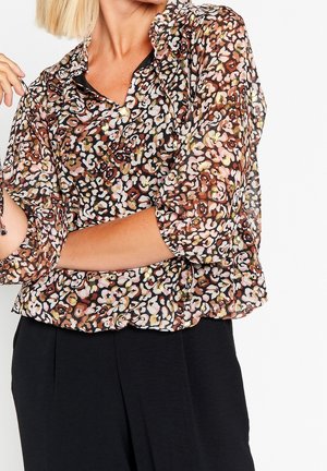 Žena má na sobě průsvitnou blůzu s dlouhými rukávy a vícebarevným květinovým vzorem a černé kalhoty s vysokým pasem na bílém pozadí.