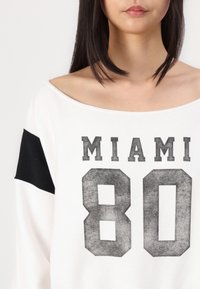 Balta sportinė palaidinė su juodais akcentais, su užrašymu "MIAMI 80" išblukusiu, ryškiu šriftu per priekinę dalį; platus viršutinės dalies apykaklės dizainas.