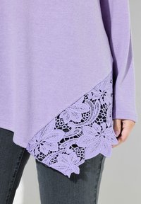 Hellblauer Pullover mit langen Ärmeln, der einen Spitzenbesatz in Lavendel mit Blumenmustern und einen asymmetrischen Saum aufweist.