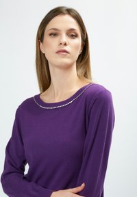 Lila Pullover mit langen Ärmeln, ausgestattet mit einem runden Halsausschnitt, der mit einer Reihe von Strasssteinen verziert ist. Weicher, glatter Stoff.