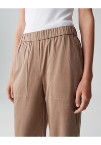 Helle braune, elastische Hosen mit zwei vorderen Taschen und einer glatten Textur. Über der Taille ist ein enganliegendes weißes Tanktop sichtbar.