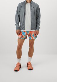 Grå lynlåsjakke, hvid t-shirt med rund hals, flerfarvede shorts med blå og orange mønstre og lyserøde sportssko med sorte detaljer.
