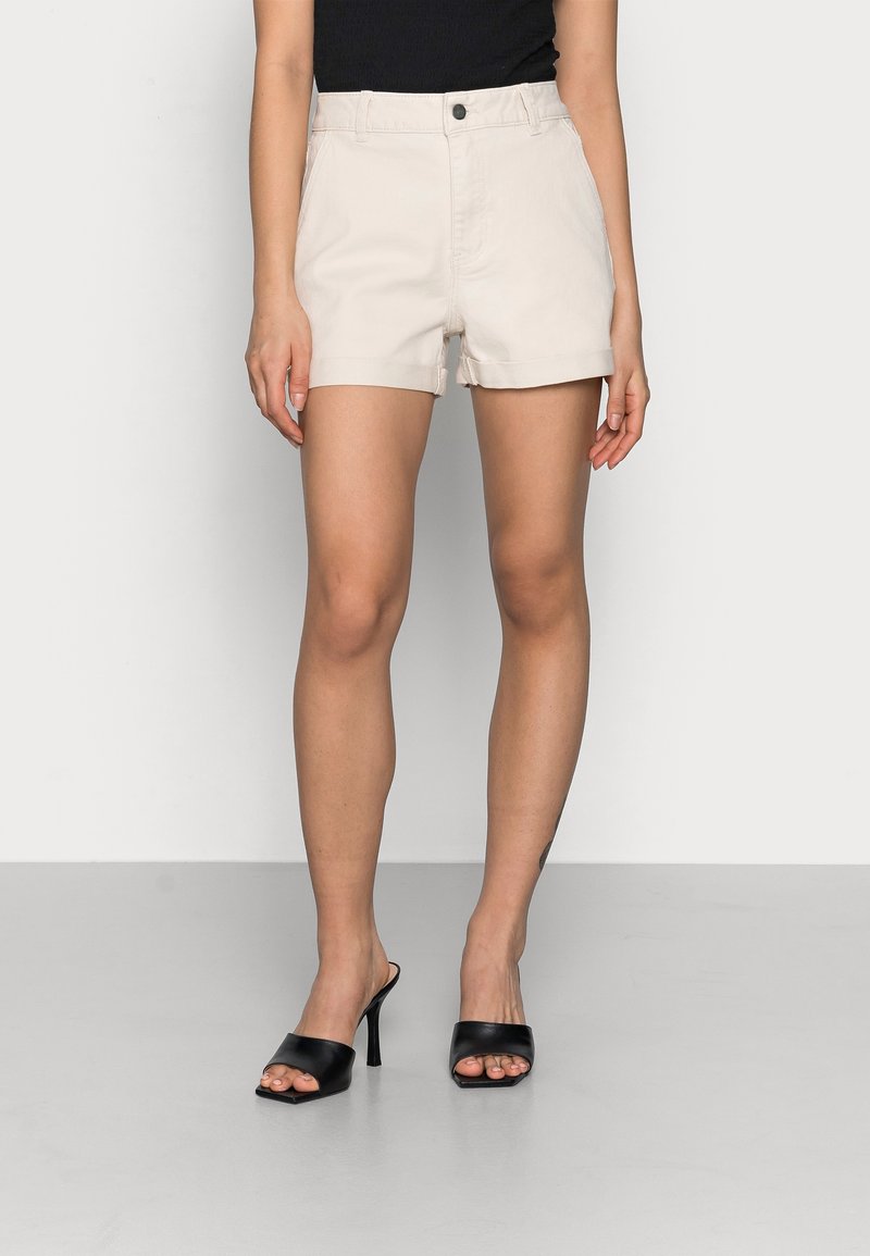 Shorts beige clair en coton avec une taille haute, dotés d'une fermeture à bouton et d'un ourlet retroussé. Associés à des sandales à talons noires à bout ouvert.