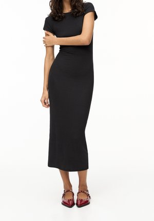 Femme portant une robe midi noire ajustée à manches courtes et des chaussures Mary Jane rouges à bout pointu avec des œillets argentés sur fond blanc.