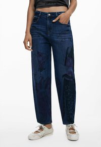 Jean en denim foncé à coupe ample avec des motifs graphiques dans des tons de violet et de noir. Comprend des poches avant et une fermeture à bouton.