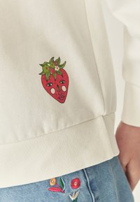 Biała bluza dresowa z kolorowym haftowanym uśmiechem truskawki z zielonymi liśćmi i kwiatami po lewej stronie, zestawiona z dżinsowymi szortami.