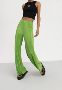 Top corto nero a costine con grafica del sole, abbinato a pantaloni larghi plissettati di un brillante verde. Indossato con sandali neri con tacco alto.