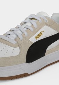 Κοντινό πλάνο σε ένα μπεζ, λευκό και μαύρο sneaker Puma με λευκά κορδόνια και καφέ σόλα, που δείχνει το χρυσό λογότυπο Puma στο πλάι.