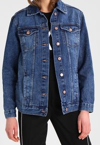 Una chaqueta de mezclilla azul con botones de cobre, bolsillos de solapa en el pecho, usada sobre una camisa negra y pantalones negros con franjas laterales blancas.