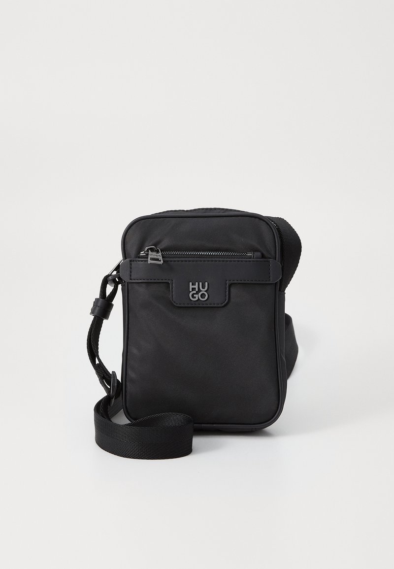Sac bandoulière noir compact avec poche avant zippée et sangle ajustable, avec logo HUGO embossé sur le rabat avant.